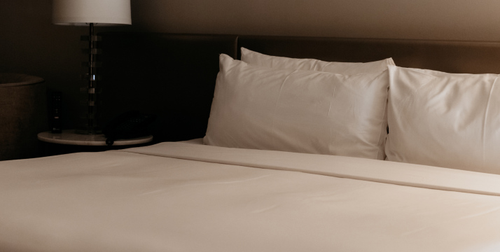 A neatly made bed with white linens and two pillows, accompanied by a bedside lamp on a small table.