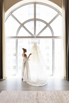 A wide room with a large arched window, a white floor, and a white rug, with a woman in a white dress standing by the window holding a wedding veil.