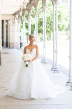 A bride in a white wedding dress stands under a covered walkway with white columns.