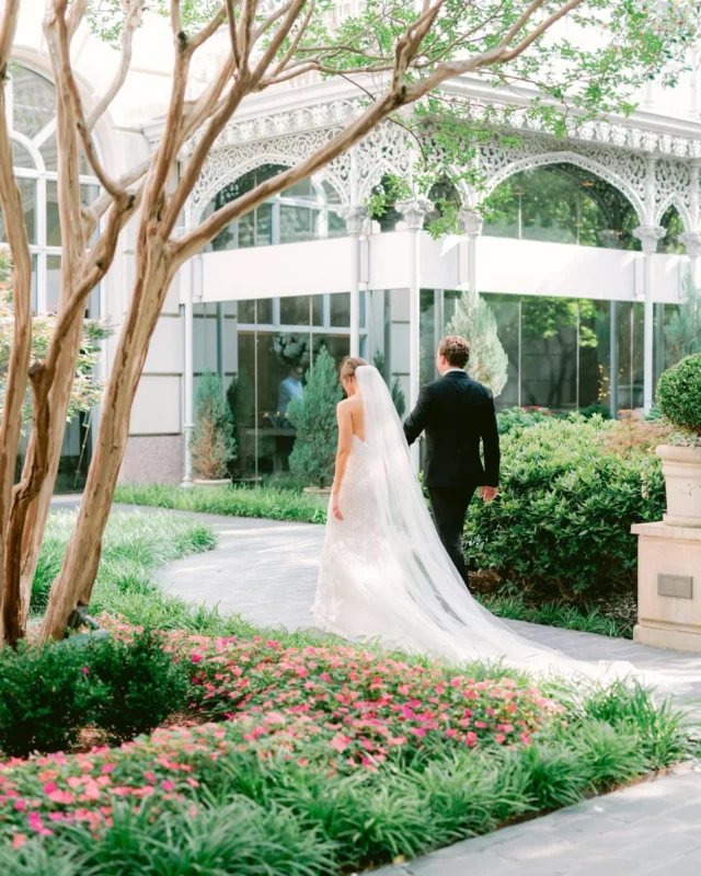 A bride and groom walk hand in hand through a beautiful garden with a greenhouse in the background.